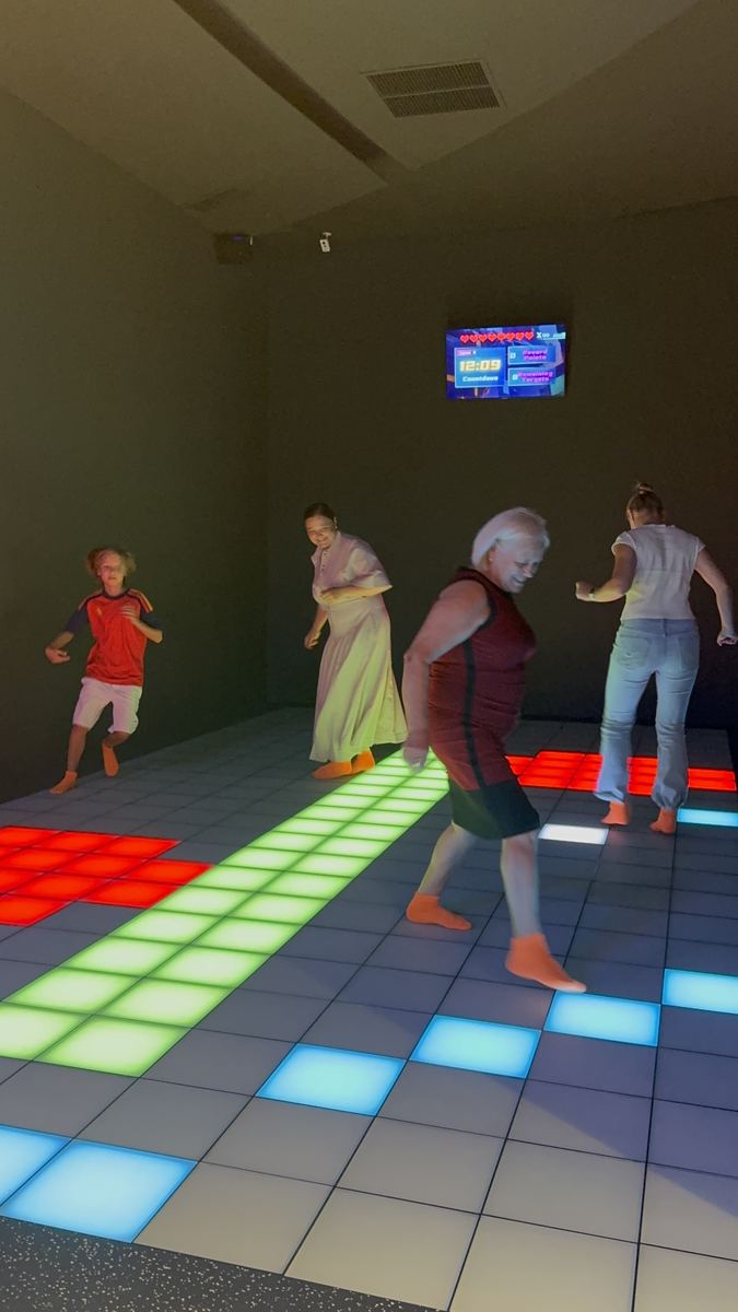 Adulți jucând pe podeaua LED verde și albă la Hai Pixel, un jucător întinzându-se după o celulă